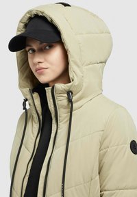 Bēšas pildītas jakas ar kapuci, kas rotāta ar melnām auklām un apaļu logotipa uzšuvi. Zem kapuces valkāta melna cepure.