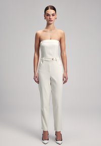 Jumpsuit bianco senza spalline con vestibilità sartoriale, tasche laterali e bottone frontale. Tessuto morbido con silhouette su misura, abbinato a tacchi a punta.