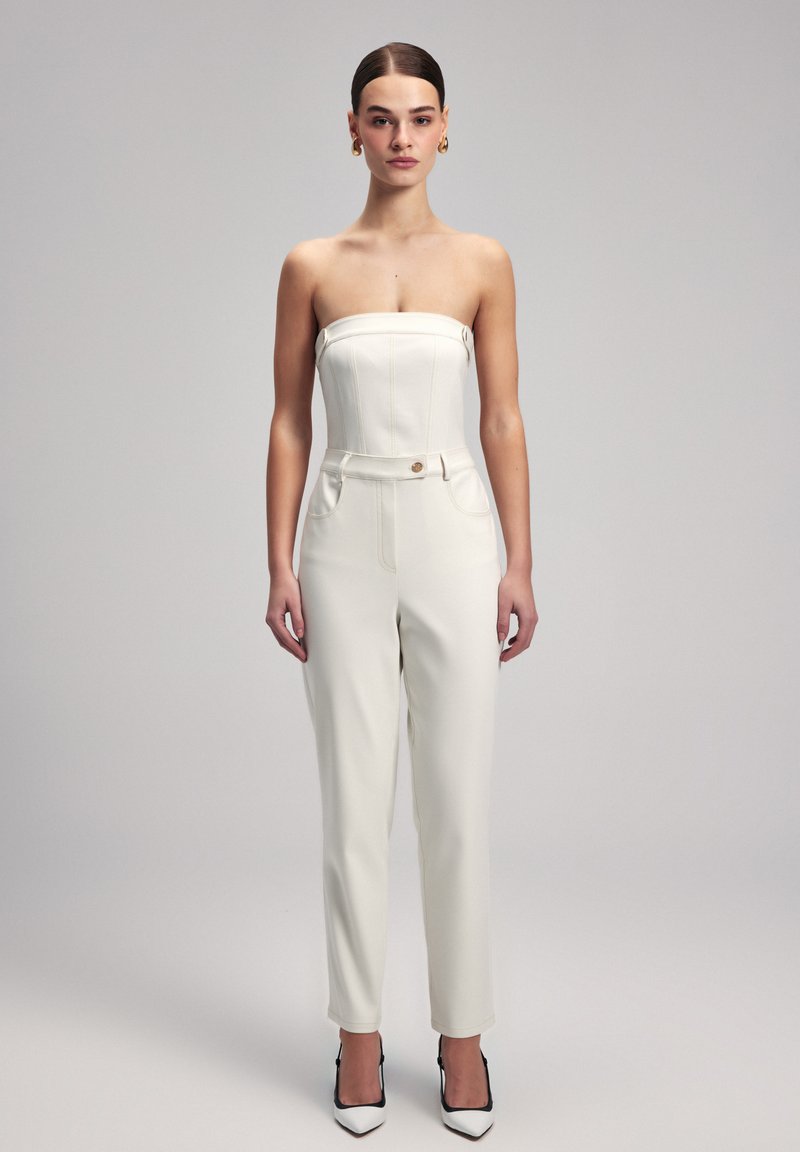 Weißer trägerloser Jumpsuit mit taillierter Passform, seitlichen Taschen und einer Vorderknopf. Geschmeidige Textur mit maßgeschneiderter Silhouette, kombiniert mit spitzen Absätzen.