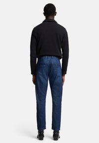 Jeans in denim blu scuro con vestibilità dritta, dotati di due tasche posteriori e un dettaglio di cucitura a contrasto. Indossati con una camicia nera.