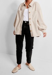 Béžová košile na knoflíky s dvěma náprsními kapsami, černé džíny s vysokým pasem a světle barevné slip-on boty, na jednobarevném pozadí.
