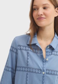 Chemise bleu clair avec un col boutonné, présentant une broderie à œillets en motif quadrillé, et de petits boutons blancs sur le devant.
