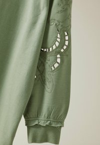 Sudadera de algodón verde con diseño bordado y acentos con cortes en las mangas, y un dobladillo con volantes en los puños. Textura suave.