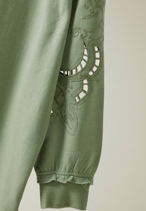 Sweatshirt en coton vert avec un design brodé, des accents découpés sur les manches et un ourlet volanté au niveau des poignets. Texture douce.