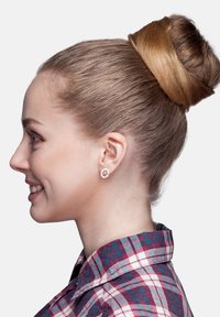 Runda, silverfärgade örhängen med små, klara stenar i en blommig design. Håret stylat i en slät, hög knut. Röd och blå rutigt skjorta.