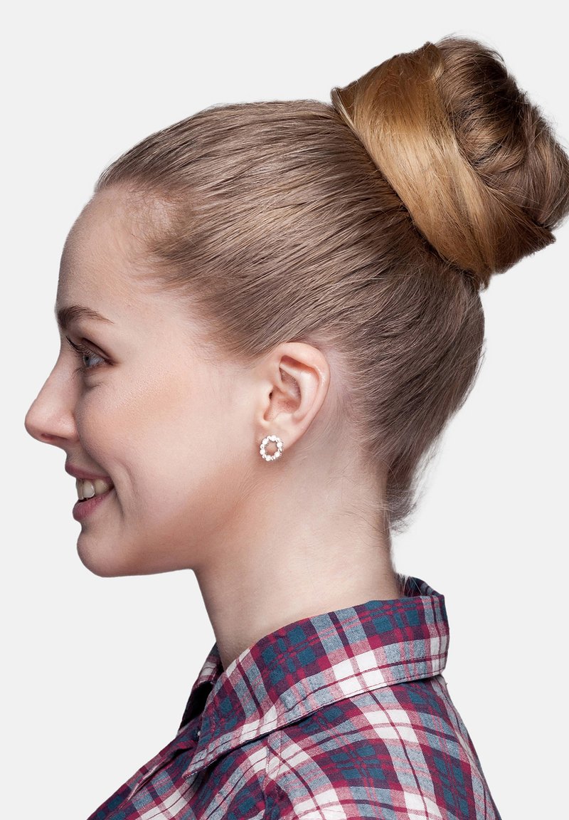 Runda, silverfärgade örhängen med små, klara stenar i en blommig design. Håret stylat i en slät, hög knut. Röd och blå rutigt skjorta.