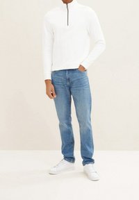 Sudadera blanca con cierre y textura acanalada, combinada con jeans de mezclilla azul claro. Los zapatos son zapatillas blancas. El fondo es neutro.