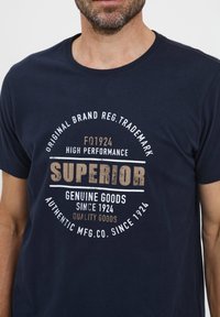 Marineblå bomulds t-shirt med rund halsudskæring, der har "SUPERIOR" tekst i stort præg og yderligere brandingdetaljer i hvid og beige.