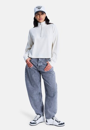 Pull-over blanc zippé avec texture côtelée, associé à un jean ample gris. Le modèle porte des baskets noires et blanches, ainsi qu'une casquette grise Roxy.