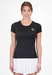 Mujer joven sonriendo, vestida con una camiseta negra ajustada de manga corta y una falda blanca plisada, frente a un fondo liso.