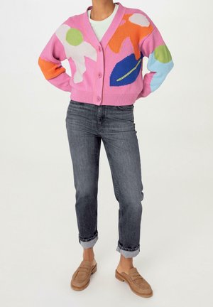 Personne portant un cardigan rose boutonné avec des formes abstraites colorées, un jean gris foncé retroussé et des mocassins marron clair sans lacets, debout les mains sur les hanches.