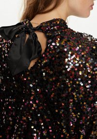 Top nero con paillettes e un fiocco al collo. Le paillettes sono multicolori, con sfumature di rosa, oro e argento, creando un aspetto texturizzato.
