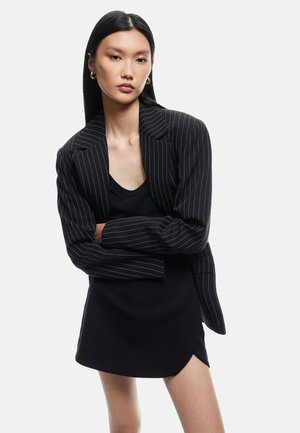 Blazer nero a righe con un design strutturato e rever a punta, indossato su una maglietta nera aderente e una gonna nera leggermente svasata.