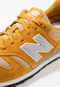 Baskets en daim jaune avec des accents en maille grise, arborant un grand logo "N" blanc et des lacets orange, sur une semelle en caoutchouc noire.