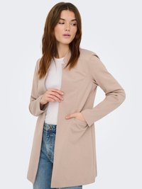 Veste beige à manches longues avec une coupe ajustée, devant ouvert et poches latérales, associée à un haut blanc et un jean en denim bleu clair.