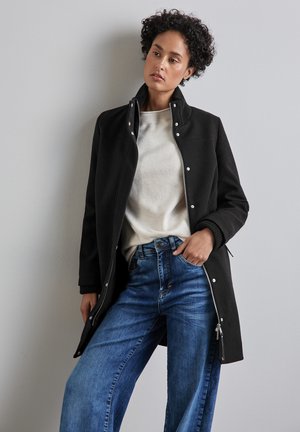 Manteau noir à fermeture éclair sur le devant avec un col haut, doté de boutons-pression, de poignets côtelés et de poches latérales, porté sur un haut crème et un jean bleu.