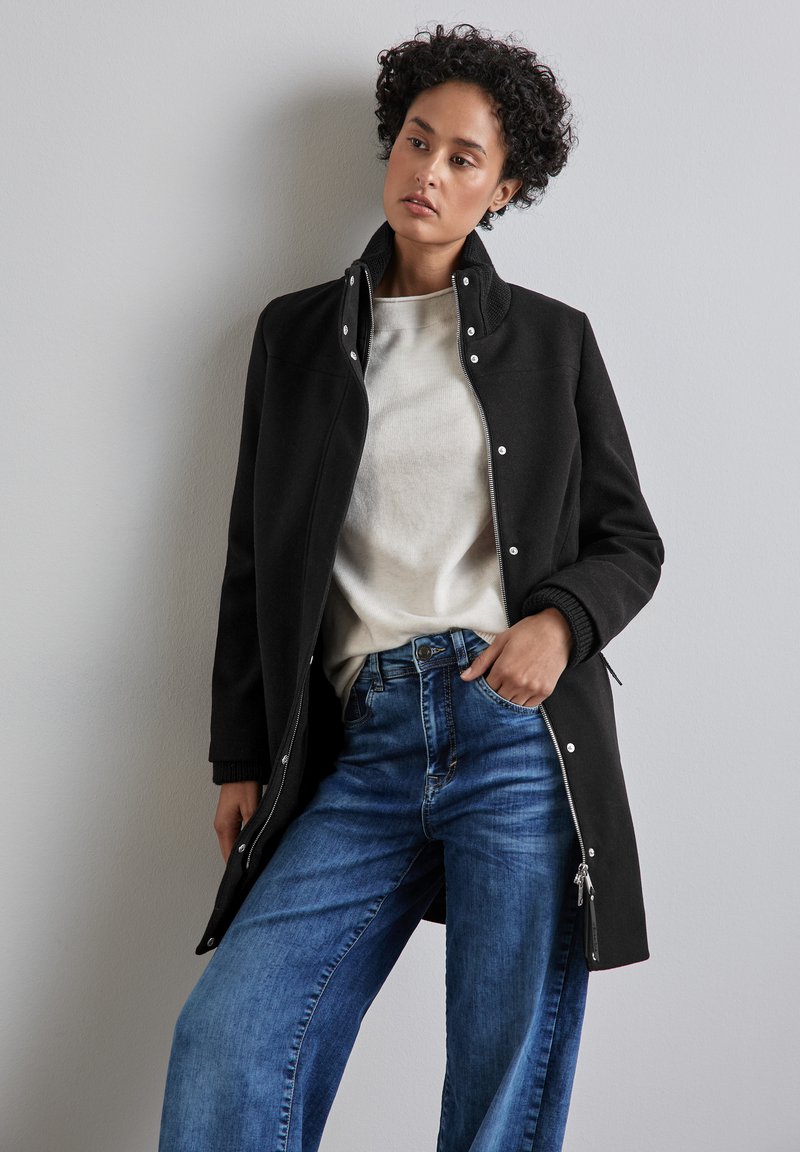 Svart zip-front jacka med hög krage, har tryckknappar, ribbade ärmmuddar och sidofickor, bärs över en krämfärgad topp och blå jeans.