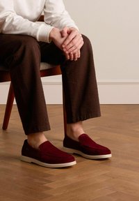 Burgundy suede slip-on shoes with white rubber soles, featuring a smooth texture and stitched detailing, worn with dark textured pants.
