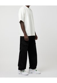 Hombre vestido con una camiseta blanca de gran tamaño, pantalones cargo negros con grandes bolsillos y zapatillas blancas de corte bajo con acentos verdes, de pie sobre un fondo blanco.