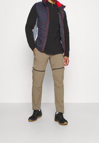 Pánský outdoorový outfit s námořnickým zatepleným vestou s červenými detaily a černým tričkem s dlouhým rukávem, doplněný béžovými kalhotami se zipem a černými botami.
