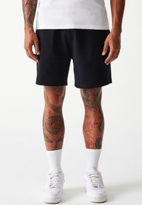 Pantaloni corti neri realizzati in tessuto morbido, con vestibilità rilassata. Indossati con sneakers bianche e calzini bianchi fino a metà polpaccio, che mostrano colorati tatuaggi sulle gambe.