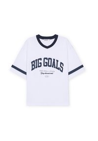 Camiseta blanca de gran tamaño con cuello en V azul marino y acentos en las mangas. Presenta el texto "GRANDES METAS" y "Y PEQUEÑOS PASOS" en impresión en negrita.