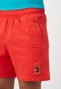 Red cotton shorts with elastic waistband, featuring a side pocket and Nike logo in a color block design on the left leg.