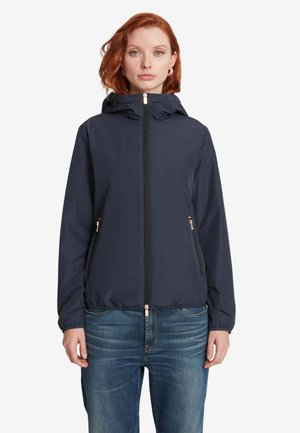 Donna con capelli rossi che indossa una giacca con cappuccio blu navy con cerniere dorate e jeans blu, in piedi davanti a uno sfondo bianco uniforme.