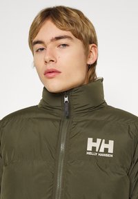 Alyvuogių spalvos pūkinė striukė su aukštu apykakle, priekiniais užtrauktukais ir Helly Hansen logotipu. Medžiaga atrodo lygi ir paminkštinta.