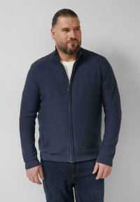 Marineblauwe trui met ritssluiting en getextureerde details op de schouders, geribde manchetten en zoom, gemaakt van een zachte stof. Gecombineerd met een donkere jeans.