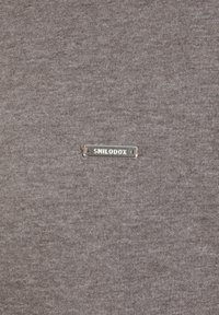 Tissu marron avec une surface lisse et texturée présentant une étiquette en métal argenté embossée avec "SMILODOX" fixée sur le côté.