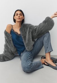 Gray faux fur coat drapes over a blue lace-trimmed camisole, paired with light denim jeans and black embellished flats.