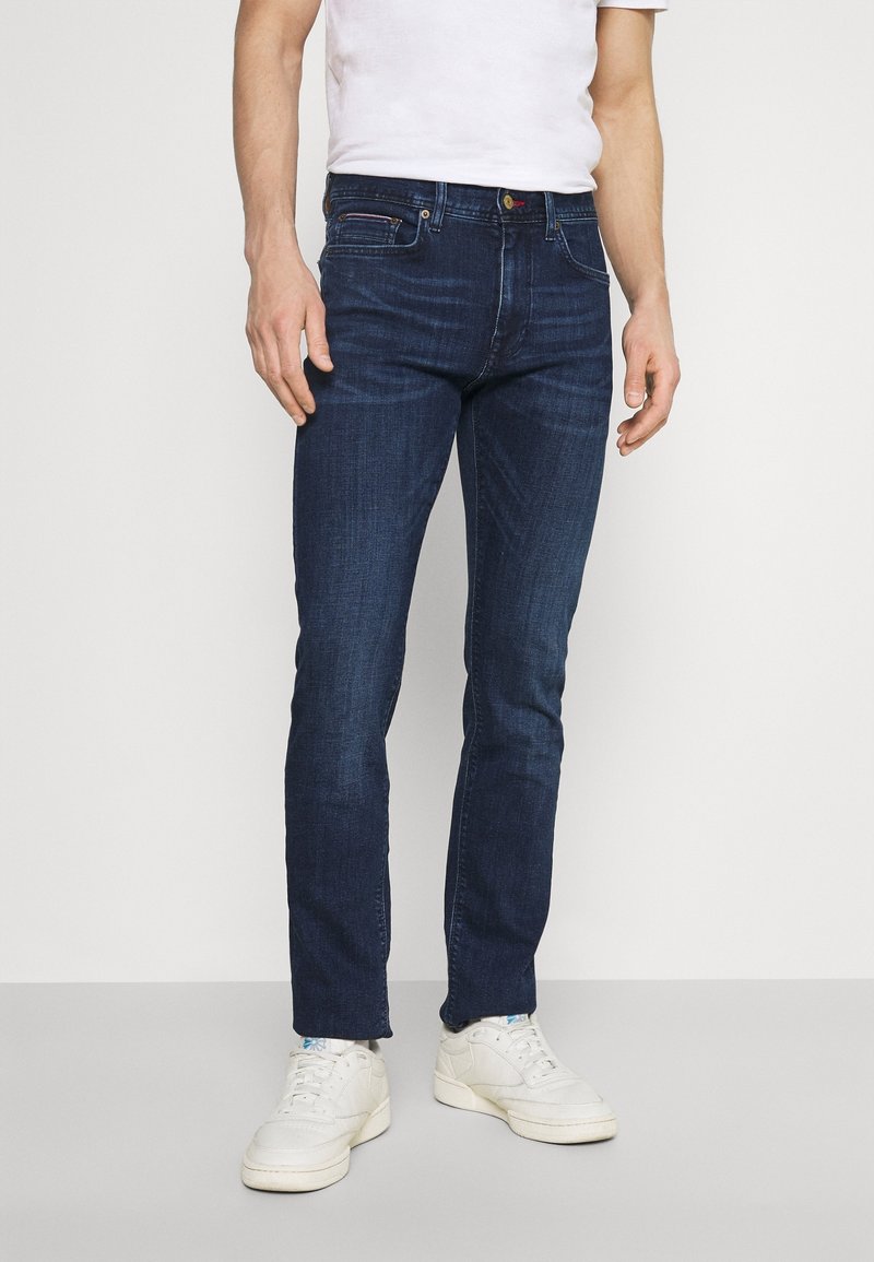 Mörkblå jeans uppvisade på en modell, tillverkade av denimtyg. Har en slim fit, synlig sömnad och en klassisk femfickdesign.