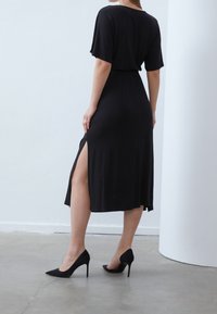 Robe midi noire à manches courtes ; dotée d'une fente latérale et d'une ceinture élastique. Portée avec des talons aiguilles noirs, sur un fond neutre.