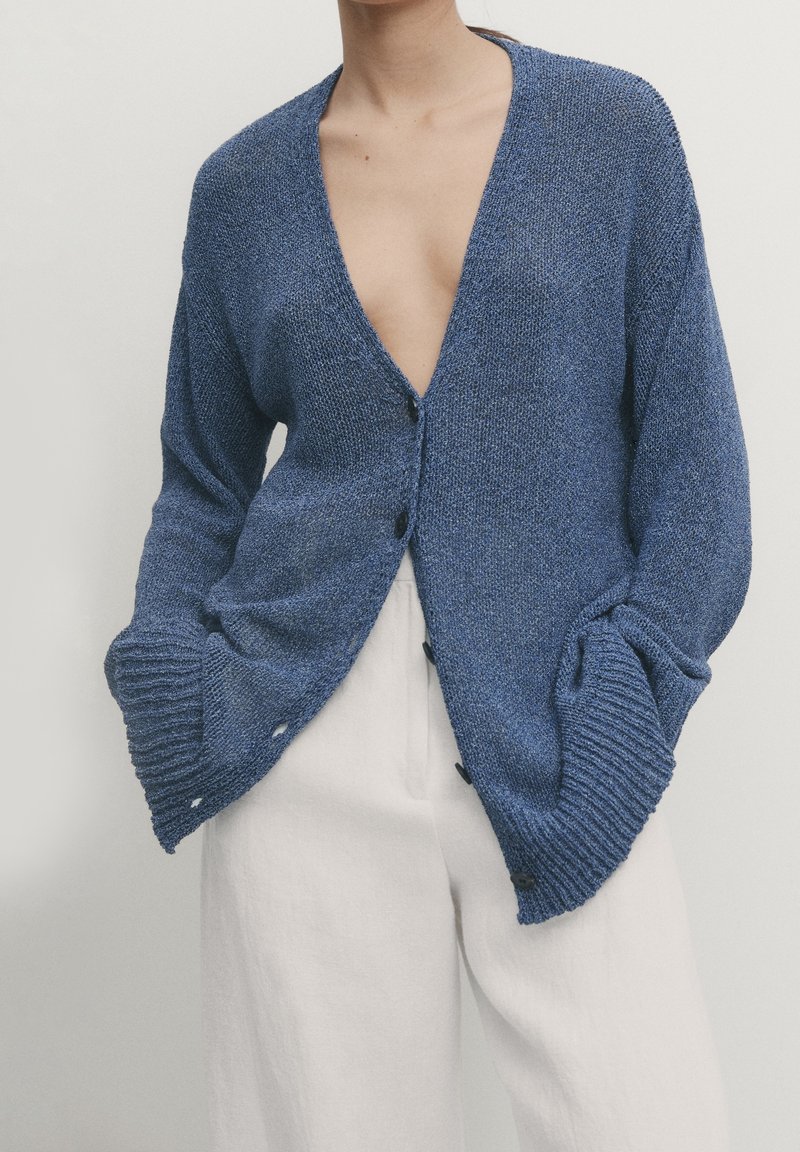 Blå stickad kofta med djup V-ringning, lång ärm, framknappning och ribbade cuffs, kompletterad med ett strukturerat tyg.