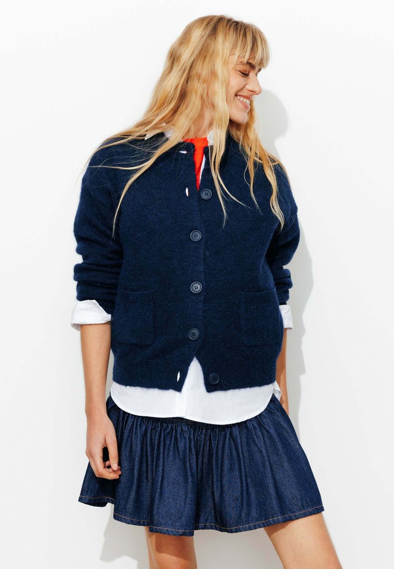 Cardigan en tricot bleu marine avec des boutons et des poches, superposé à une chemise blanche et associé à une jupe en denim foncé avec une taille froncée.