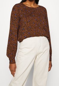 Brun blommig blus med långa ärmar, med ett texturerat mönster av orangea blad och blå blommor, tillsammans med vita byxor.