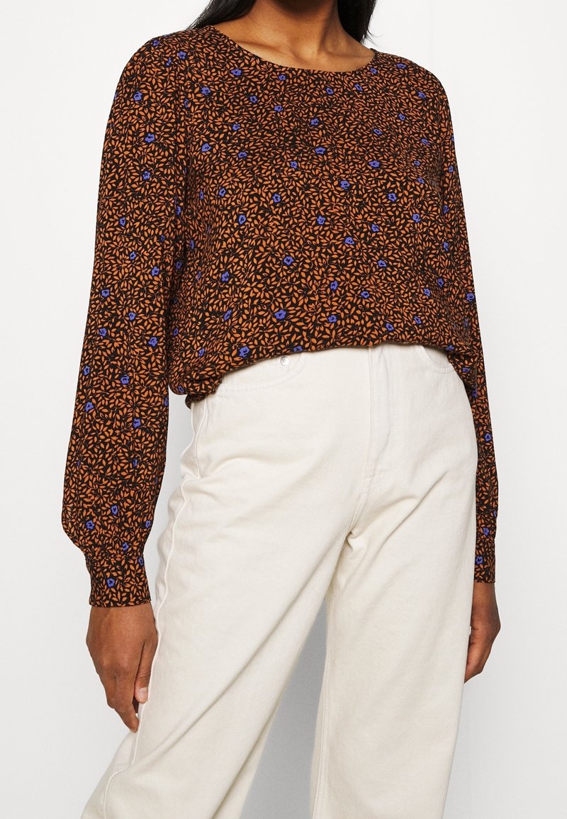 Brun blommig blus med långa ärmar, med ett texturerat mönster av orangea blad och blå blommor, tillsammans med vita byxor.