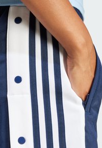 Tamnoplave sportske kratke hlače s bijelim trakama, s detaljem na kopčanje i bočnim džepovima. Tkanina izgleda glatko i teksturirano.