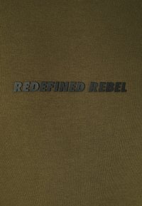 Olivgrüner Stoff mit dem Text "REDEFINED REBEL" in fettem, schwarzem Schriftzug. Glatte Textur mit einem gerippten Muster.