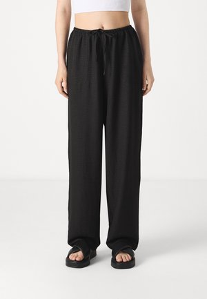 Persoon die losse zwarte broek met trekkoord en zwarte sandalen draagt, staand tegen een witte achtergrond, zichtbaar van de taille tot de voeten.