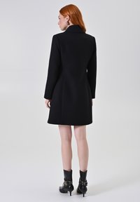 Cappotto nero su misura con un design strutturato, maniche lunghe e colletto alto. Texture liscia e lunghezza al ginocchio. Abbinato a stivaletti neri alla caviglia.