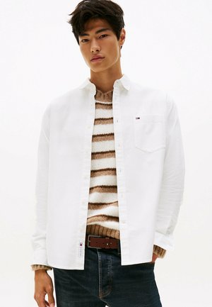 Jonge man in een wit overhemd over een bruin-wit gestreepte trui, gecombineerd met donkere jeans en een leren bruine riem.