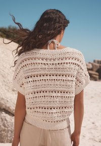 Witte gehaakte top met een open breipatroon en een geribbelde zoom. Bevat korte mouwen en een halterkoord aan de achterkant. Gepaard met lichtbeige broeken.