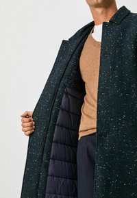 Mörkgrön tweedjacka med en herringbone-mönster, med en quiltad marinblå foder och enstaka knappstängning. Brun stickad tröja under.