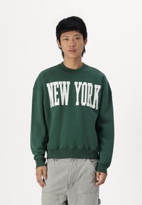 Green sweatshirt with "NEW YORK" printed in white across the chest. Features a crew neck and ribbed cuffs. Smooth fabric texture.