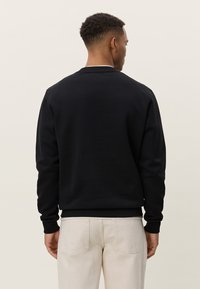 Μαύρο φούτερ με στρογγυλή λαιμόκοψη και ριμπ μανσέτες, με λεία υφή και απλό σχέδιο. Φοριέται με ανοιχτόχρωμο παντελόνι.