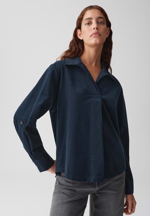 Frau mit welligem braunem Haar, die ein dunkelblaues Langarmhemd mit Kragen und graue Jeans trägt, steht vor einem schlichten hellen Hintergrund.