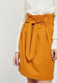 Jupe moutarde à taille haute, équipée d'une ceinture nouée. Le tissu présente une texture lisse et est conçu avec des plis à l'avant pour plus d'ampleur.