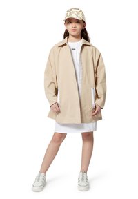 Chaqueta ligera color beige con un ajuste relajado, bolsillos con cremallera blanca, usada sobre un vestido blanco. El modelo lleva una gorra con estampado y zapatillas blancas.
